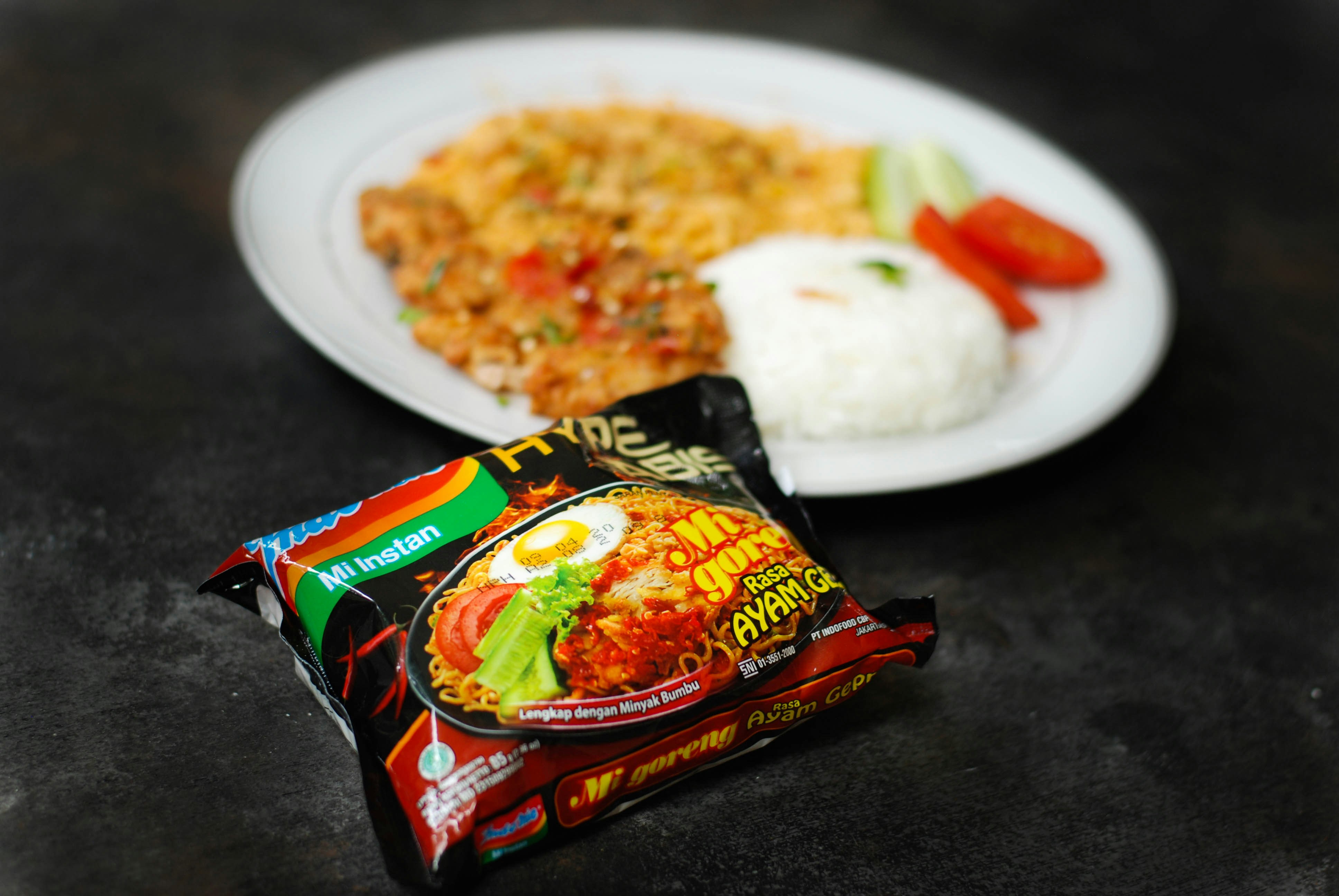 A packet of instant noodles is in the foreground, featuring colorful packaging with an image of the prepared noodles and labeled with text. In the background, there is a white plate containing cooked rice, slices of tomato and cucumber, along with another dish that appears to be seasoned and cooked with herbs and vegetables.