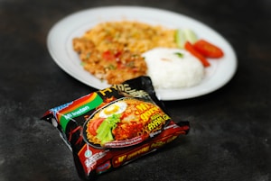 A packet of instant noodles is in the foreground, featuring colorful packaging with an image of the prepared noodles and labeled with text. In the background, there is a white plate containing cooked rice, slices of tomato and cucumber, along with another dish that appears to be seasoned and cooked with herbs and vegetables.