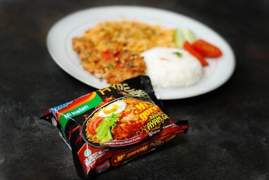 A packet of instant noodles is in the foreground, featuring colorful packaging with an image of the prepared noodles and labeled with text. In the background, there is a white plate containing cooked rice, slices of tomato and cucumber, along with another dish that appears to be seasoned and cooked with herbs and vegetables.