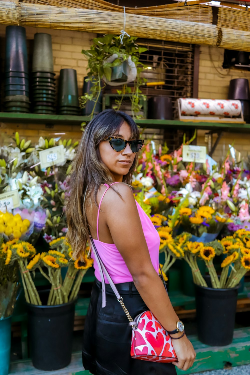 Femme en débardeur rose portant des lunettes de soleil noires debout près de fleurs