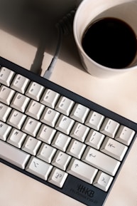 Close-up of hands typing on a minimalist keyboard beside a cup of coffee and a notebook.