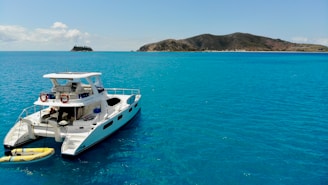 A luxurious catamaran is anchored on a calm, turquoise sea. The clear blue water gently ripples around the boat, and a small inflatable raft is attached to the stern. In the distance, a hilly island is visible under a bright blue sky with a few scattered clouds.