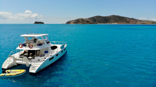 A luxurious catamaran is anchored on a calm, turquoise sea. The clear blue water gently ripples around the boat, and a small inflatable raft is attached to the stern. In the distance, a hilly island is visible under a bright blue sky with a few scattered clouds.