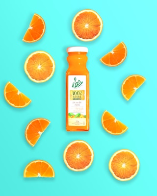 A bottle of orange juice is centrally placed among various orange slices on a bright turquoise background. The label on the bottle indicates it is 100% natural orange juice.
