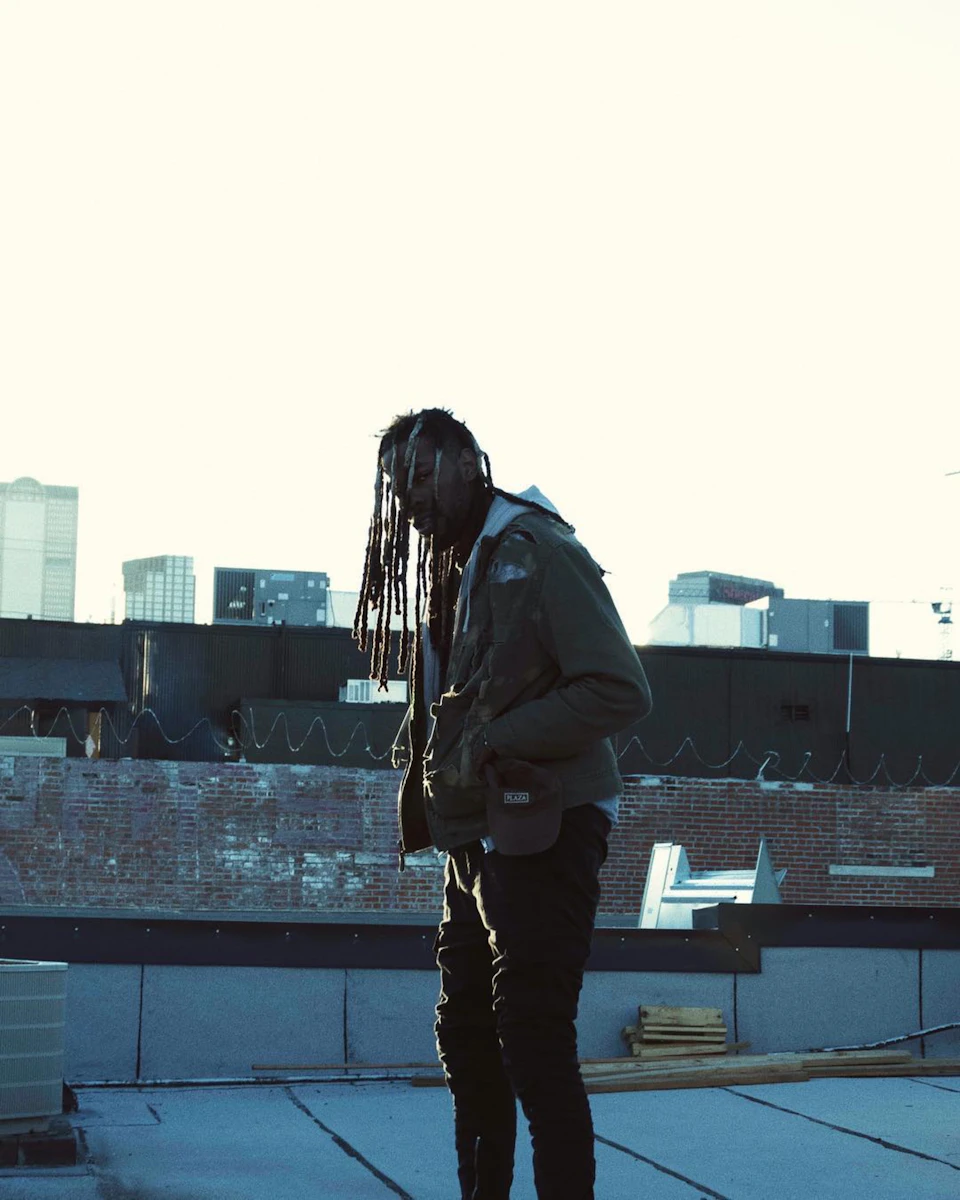 Person with dreadlocks stands on a rooftop, wearing clothing and holding a bag