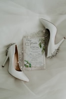 A pair of elegant white high-heeled shoes are placed alongside a wedding invitation. The invitation is adorned with delicate floral and leaf designs, accompanied by small decorative pearls and beads.