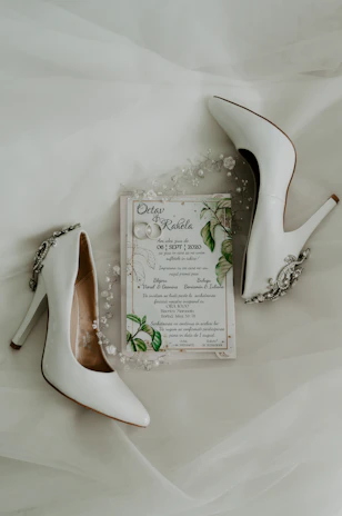 A pair of elegant white high-heeled shoes are placed alongside a wedding invitation. The invitation is adorned with delicate floral and leaf designs, accompanied by small decorative pearls and beads.