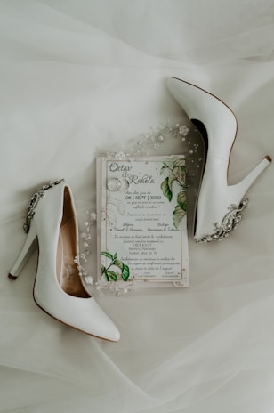 A pair of elegant white high-heeled shoes are placed alongside a wedding invitation. The invitation is adorned with delicate floral and leaf designs, accompanied by small decorative pearls and beads.