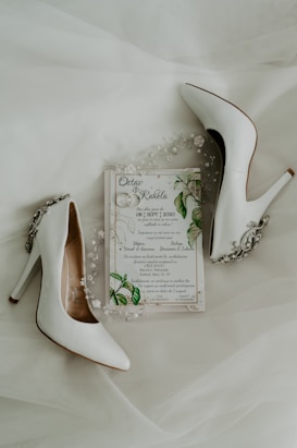 A pair of elegant white high-heeled shoes are placed alongside a wedding invitation. The invitation is adorned with delicate floral and leaf designs, accompanied by small decorative pearls and beads.