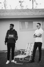 Two men stand outdoors near a house with a window, a roof, and a lawn. One wears a hoodie labeled 'FAKE' and holds a phone. The other wears sunglasses and a buttoned shirt. They are next to a pair of outdoor chairs on a grassy area.