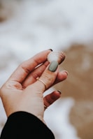 A hand holding a small seashell between fingers. The hand has neatly manicured nails with a muted green polish on the thumb and rings on several fingers. The background is blurred, suggesting a sandy beach setting.