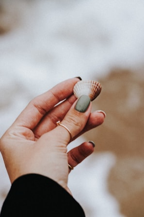 A hand holding a small seashell between fingers. The hand has neatly manicured nails with a muted green polish on the thumb and rings on several fingers. The background is blurred, suggesting a sandy beach setting.
