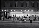 A bustling street scene at night with people walking along the sidewalk. A neon sign reads 'Kino Kultura' above a cinema or theater entrance, and a restaurant named 'Cinema Paradiso' is visible. A man rides a scooter in the foreground, while groups of people stroll casually in various directions.