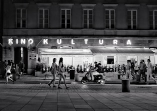 A bustling street scene at night with people walking along the sidewalk. A neon sign reads 'Kino Kultura' above a cinema or theater entrance, and a restaurant named 'Cinema Paradiso' is visible. A man rides a scooter in the foreground, while groups of people stroll casually in various directions.