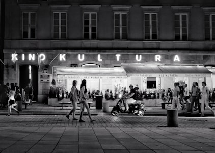 A bustling street scene at night with people walking along the sidewalk. A neon sign reads 'Kino Kultura' above a cinema or theater entrance, and a restaurant named 'Cinema Paradiso' is visible. A man rides a scooter in the foreground, while groups of people stroll casually in various directions.