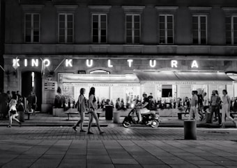 A bustling street scene at night with people walking along the sidewalk. A neon sign reads 'Kino Kultura' above a cinema or theater entrance, and a restaurant named 'Cinema Paradiso' is visible. A man rides a scooter in the foreground, while groups of people stroll casually in various directions.