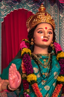 A beautifully adorned statue of a deity featuring a serene expression and intricate jewelry. The figure is draped in a turquoise sari with gold accents and wears a golden crown. Vibrant flowers in shades of yellow and magenta are arranged around the neck, adding to the regal appearance. The right hand is raised in a symbolic gesture.