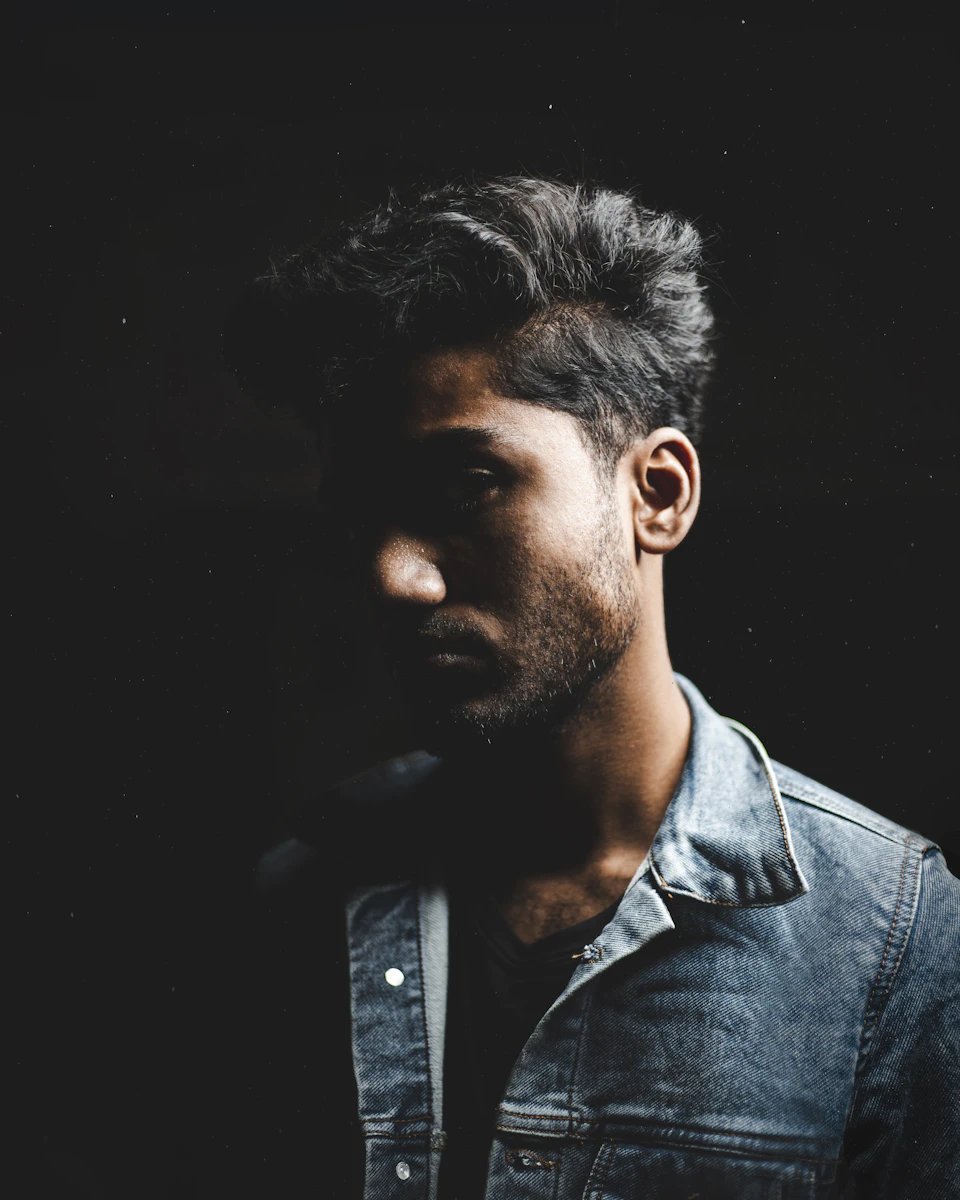 Man stands against black background wearing blue denim jacket with partially obscured face