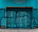 A large corrugated metal garage door or shutter painted in teal, covered in graffiti. The wall and garage door have a weathered texture, and the area surrounding the door is painted to blend with the same color scheme.