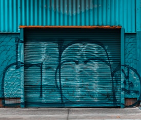 A large corrugated metal garage door or shutter painted in teal, covered in graffiti. The wall and garage door have a weathered texture, and the area surrounding the door is painted to blend with the same color scheme.