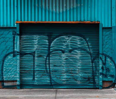 A large corrugated metal garage door or shutter painted in teal, covered in graffiti. The wall and garage door have a weathered texture, and the area surrounding the door is painted to blend with the same color scheme.
