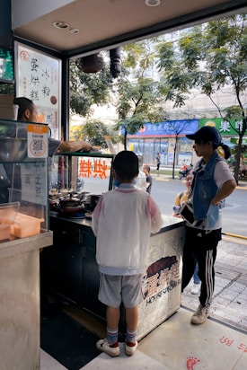 A street food stall is set up in an urban environment with a man cooking behind the counter. A young boy is waiting to be served, standing in front of the stall with a menu on display. Another individual casually stands nearby, possibly waiting or chatting. The scene captures a casual and lively street atmosphere, with trees and shops visible in the background. Signs and advertisements in various languages are posted around, adding to the bustling city vibe.