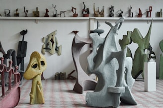 A room filled with various abstract sculptures made of different materials and colors. Larger sculptures dominate the foreground, with smaller pieces on shelves in the background. The sculptures feature unique shapes and figures, combining smooth and jagged elements.