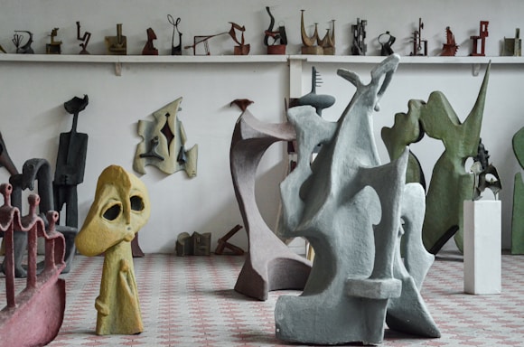 A room filled with various abstract sculptures made of different materials and colors. Larger sculptures dominate the foreground, with smaller pieces on shelves in the background. The sculptures feature unique shapes and figures, combining smooth and jagged elements.