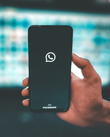 A hand is holding a smartphone displaying the WhatsApp logo on its screen. The text 'from Facebook' appears below the logo. The background is blurred, with cool blue tones and a grid-like pattern.