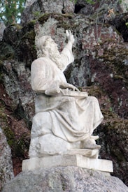 A carved stone statue of a bearded figure sits against a rocky background. The figure is draped in robes and appears to be gesturing upward with one hand while holding a tablet or book in the other. The setting is natural, with moss-covered rocks and some greenery visible.