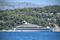 A large, luxurious yacht is anchored near a forested coastline with dense green trees. The backdrop features rugged, rocky mountains under a clear, blue sky. The yacht is modern and sleek, predominantly white with multiple decks.