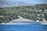 A large, luxurious yacht is anchored near a forested coastline with dense green trees. The backdrop features rugged, rocky mountains under a clear, blue sky. The yacht is modern and sleek, predominantly white with multiple decks.