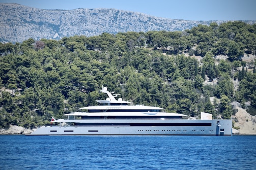 A large, luxurious yacht is anchored near a forested coastline with dense green trees. The backdrop features rugged, rocky mountains under a clear, blue sky. The yacht is modern and sleek, predominantly white with multiple decks.