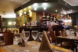 A cozy restaurant interior featuring a bar area with an array of bottles and glasses. The tables are elegantly set with wine glasses and folded napkins. Warm lighting creates an inviting atmosphere, with wooden chairs and furniture adding to the charm.