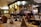 A cozy restaurant interior featuring a bar area with an array of bottles and glasses. The tables are elegantly set with wine glasses and folded napkins. Warm lighting creates an inviting atmosphere, with wooden chairs and furniture adding to the charm.