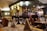 A cozy restaurant interior featuring a bar area with an array of bottles and glasses. The tables are elegantly set with wine glasses and folded napkins. Warm lighting creates an inviting atmosphere, with wooden chairs and furniture adding to the charm.