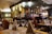 A cozy restaurant interior featuring a bar area with an array of bottles and glasses. The tables are elegantly set with wine glasses and folded napkins. Warm lighting creates an inviting atmosphere, with wooden chairs and furniture adding to the charm.