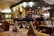 A cozy restaurant interior featuring a bar area with an array of bottles and glasses. The tables are elegantly set with wine glasses and folded napkins. Warm lighting creates an inviting atmosphere, with wooden chairs and furniture adding to the charm.