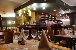 A cozy restaurant interior featuring a bar area with an array of bottles and glasses. The tables are elegantly set with wine glasses and folded napkins. Warm lighting creates an inviting atmosphere, with wooden chairs and furniture adding to the charm.