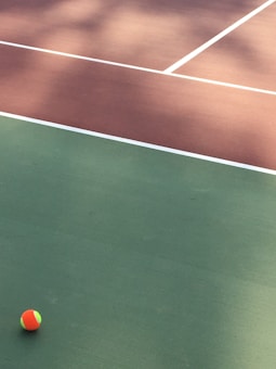A tennis court with distinct green and brown sections divided by white lines. A small, colorful tennis ball is positioned on the green surface.