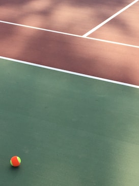 A tennis court with distinct green and brown sections divided by white lines. A small, colorful tennis ball is positioned on the green surface.