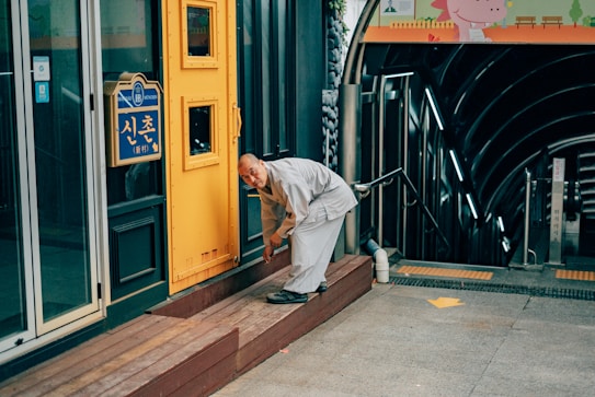 A person wearing traditional attire is bending down near a yellow and blue storefront with Korean signage. The surrounding area includes a modern staircase leading underground and a colorful sign featuring a character at the top.