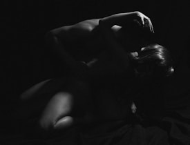 A black and white image captures two individuals embracing closely, surrounded by deep shadows. Their bodies are intertwined, creating a sense of intimacy and emotional connection.