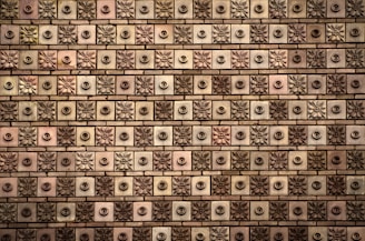 A textured wall comprised of square tiles, each featuring a floral or circular design. The tiles are arranged in a grid-like pattern, alternating between floral motifs and circular knobs. The color palette ranges from shades of brown to beige, with subtle variations in each tile.