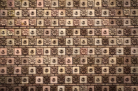 A textured wall comprised of square tiles, each featuring a floral or circular design. The tiles are arranged in a grid-like pattern, alternating between floral motifs and circular knobs. The color palette ranges from shades of brown to beige, with subtle variations in each tile.