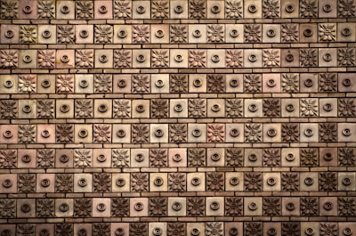 A textured wall comprised of square tiles, each featuring a floral or circular design. The tiles are arranged in a grid-like pattern, alternating between floral motifs and circular knobs. The color palette ranges from shades of brown to beige, with subtle variations in each tile.
