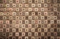 A textured wall comprised of square tiles, each featuring a floral or circular design. The tiles are arranged in a grid-like pattern, alternating between floral motifs and circular knobs. The color palette ranges from shades of brown to beige, with subtle variations in each tile.