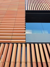 A building facade with a modern architectural design featuring vertical and horizontal lines. The facade consists of orange-brown slats and a section of reflective glass, providing a contrast in textures.