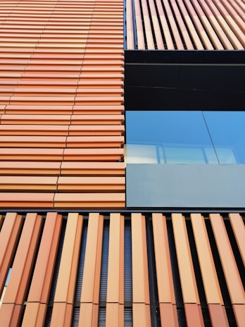 A building facade with a modern architectural design featuring vertical and horizontal lines. The facade consists of orange-brown slats and a section of reflective glass, providing a contrast in textures.