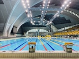 An indoor swimming pool with multiple lanes, brightly lit by overhead ceiling lights. The pool area is surrounded by large, arched structures, and an array of seats is visible on the right side. The water in the pool is clear, reflecting the bright lights above.